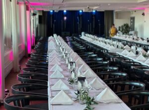 Pitkä pöytä, jossa on valkea pöytäliina ja valkeat servetit. Pöydällä on kukkakoristelut. Pöydän molemmin puolin on tuoleja.